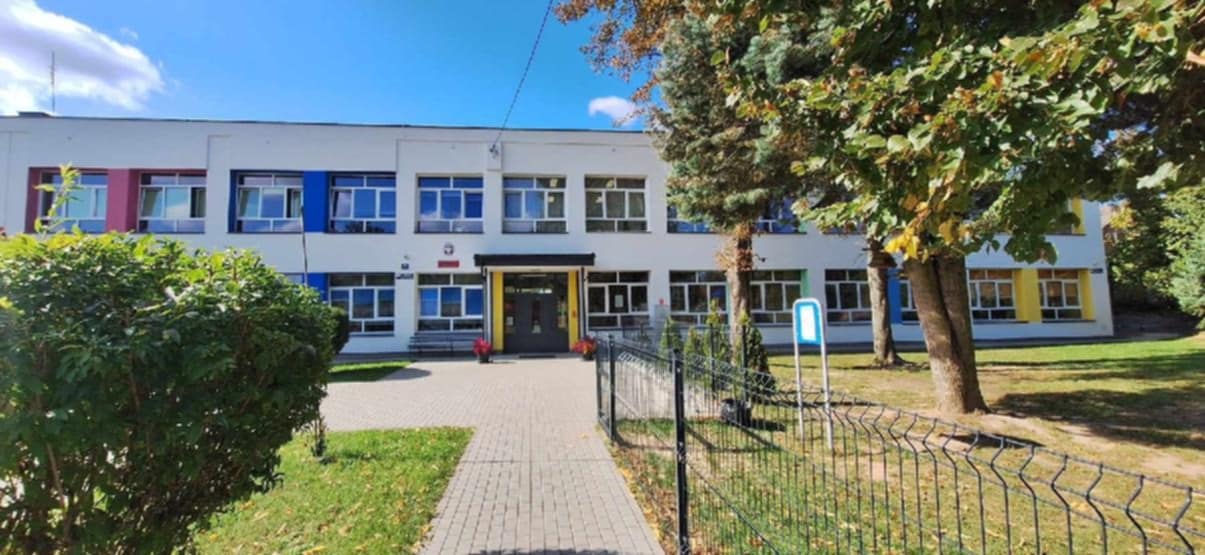 Budynek szkoły w słoneczny dzień. Dwupiętrowa biała elewacja z kolorowymi ramami okien, przed wejściem kostka brukowa, trawnik i drzewa.