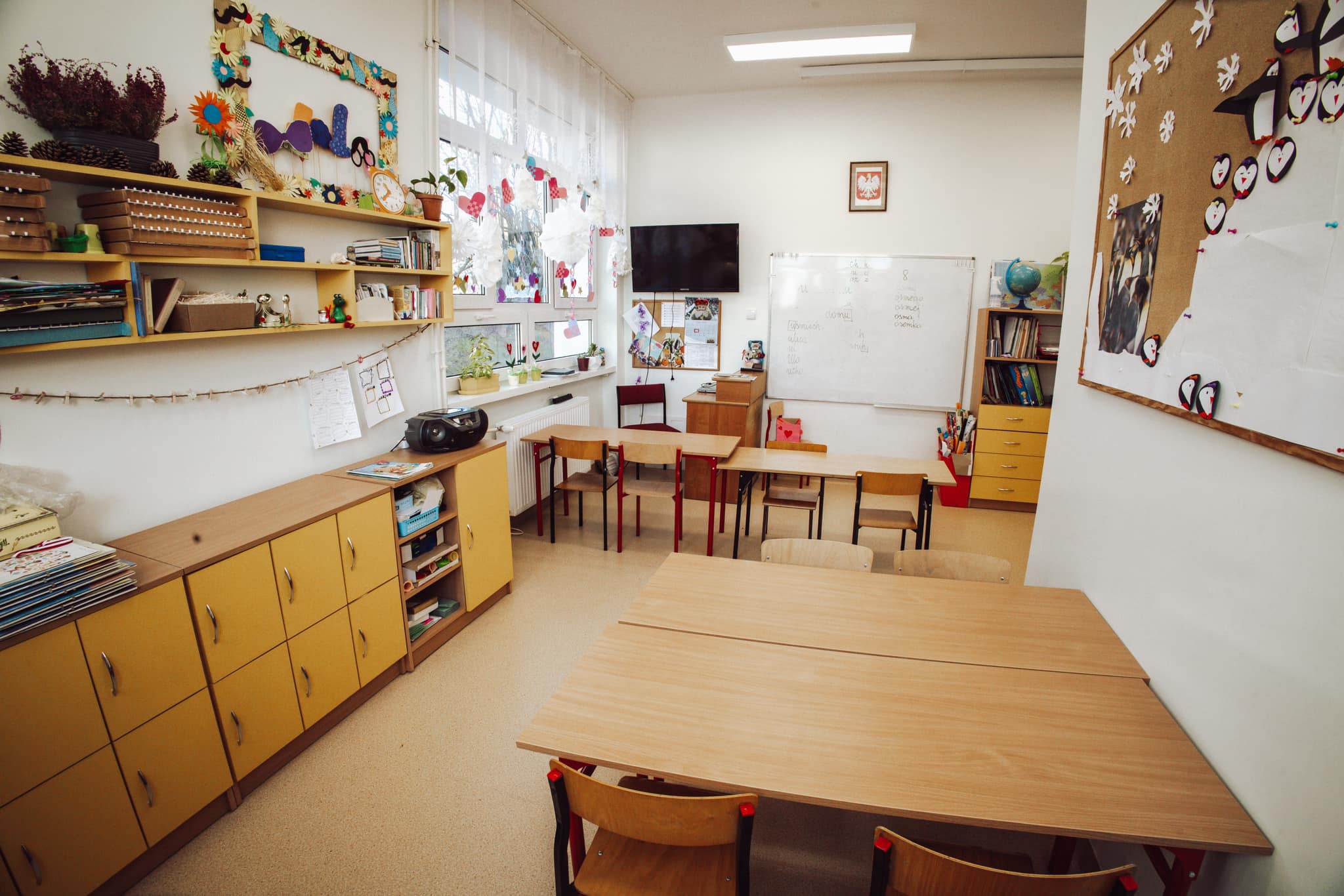 Sala lekcyjna z żółtymi szafkami, tablicą, telewizorem i pracami uczniów na ścianie. Przy oknie stoją rośliny w doniczkach.